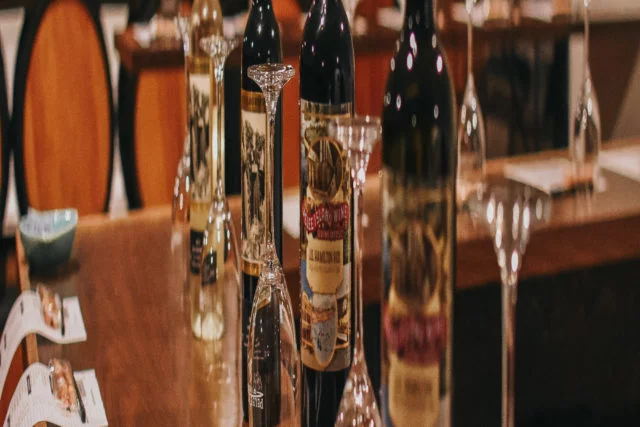 Bottles of JS Hamilton VQA from Pelee Island Winery line up with glassware on the tasting bar at the Pelee Island Winery Pavilion on Pelee Island with wine barrels in the background.