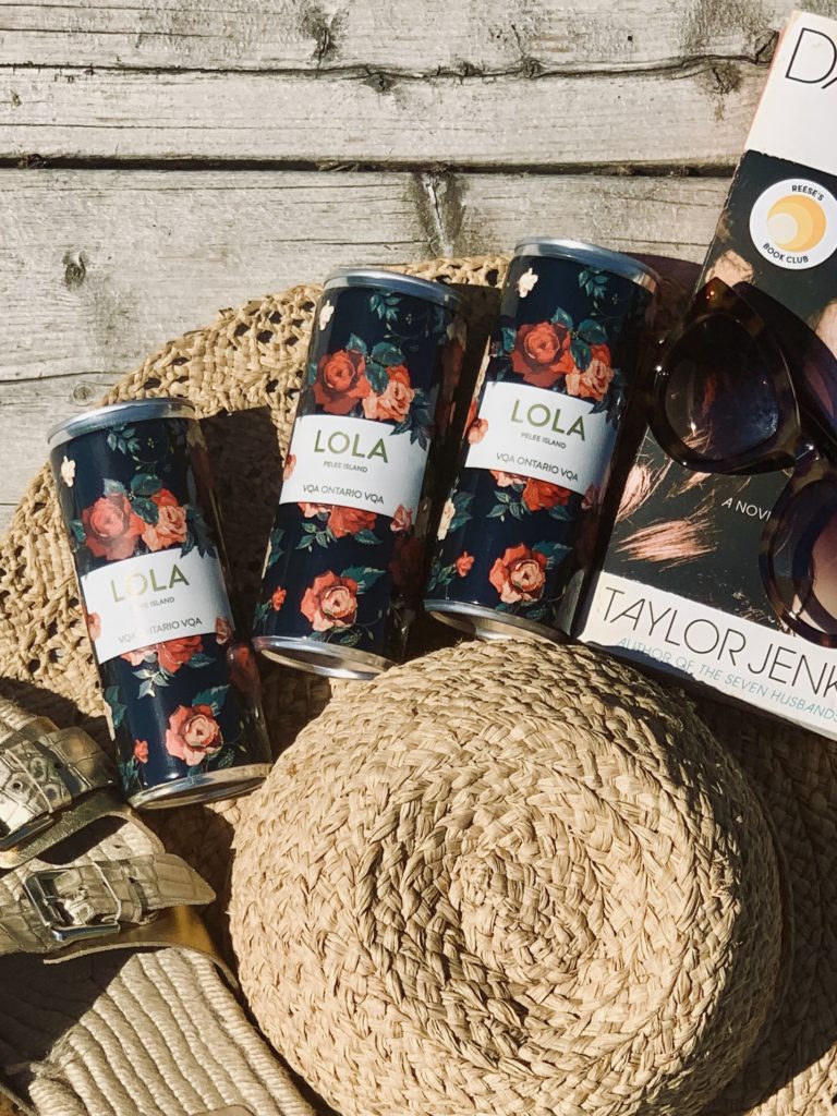 Pelee Island Winery Lola Sparkling Blush Rosé in 250 mL slim cans on deck with book, straw hat, sunglasses and sandals.