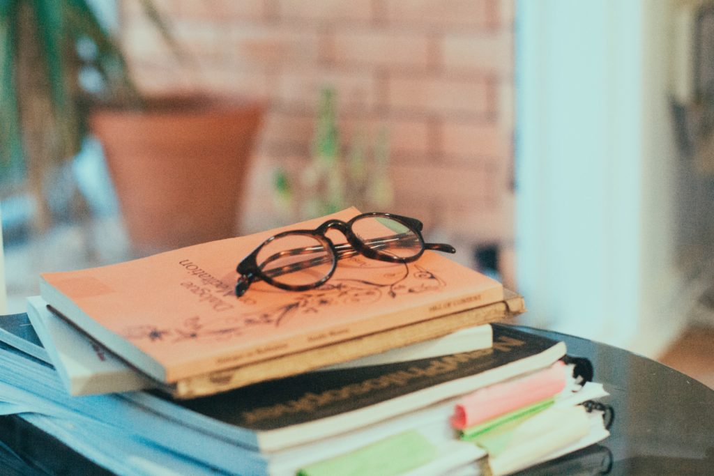 Stack of books on a table with a pair of reading glasses. 